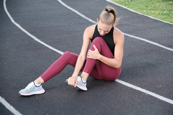 スポーツウェアの若い運動選手の女の子は、テクスチャの表面に座っており、重度の脚の痛み、痛みを伴う痙攣を持っています。彼の手で足を絞ると彼の歯をきれいに. — ストック写真