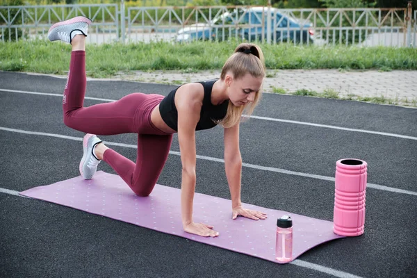 Chica de fitness haciendo ejercicio de piernas en estera de yoga en el estadio al aire libre. Ajuste mujer con fuerte cuerpo muscular en un chándal de moda haciendo ejercicios contra una pared de hormigón. — Foto de Stock