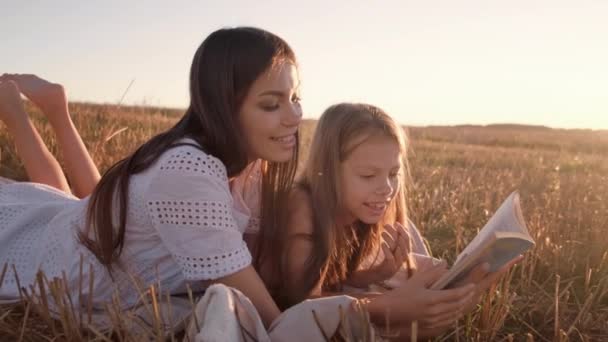 Madre e hija están tumbadas en una manta leyendo un libro y contando historias divertidas sobre el fondo de la puesta del sol — Vídeo de stock