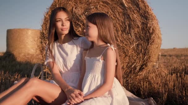 Μητέρα και κόρη με λευκό φόρεμα κάθονται σε ένα χωράφι. Η μαμά φιλάει την κόρη της στο μέτωπο και την αγκαλιάζει. — Αρχείο Βίντεο