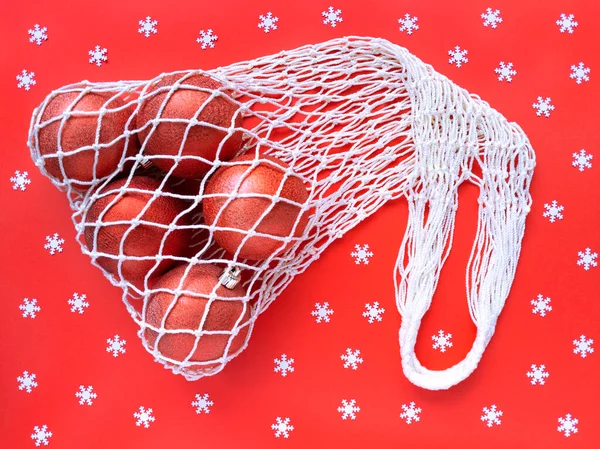 White string cotton eco bag with five christms tree balls on red background and scattered white snowflakes. Holiday simple flat lay. Ecology zero waste concept. Stock photography.