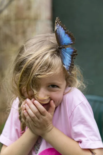 她的头发上有一只蓝色蝴蝶的傻笑女孩的肖像 中等接近 聚焦前景 — 图库照片