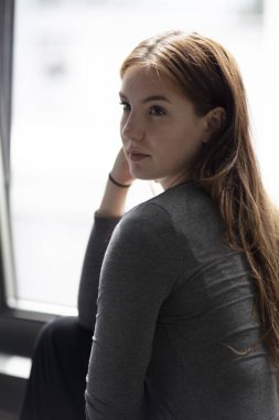 Elini çenesindeki geliyor gün ışığı alan bir pencere oturan bir genç çekici kadın güzellik portresi. Orta atış. Seçici odak. 