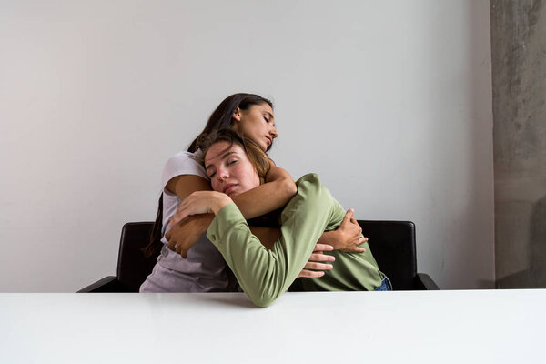 Two women performing artistic poses seated at large bureau. Loving embrace. Medium shot. Front facing. 