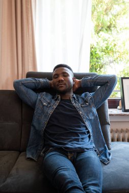 Genç siyahi adam oturma odasındaki kanepede ellerini başının arkasına koyup kestiriyor. Ön manzara. Üç çeyrek uzunluk. 