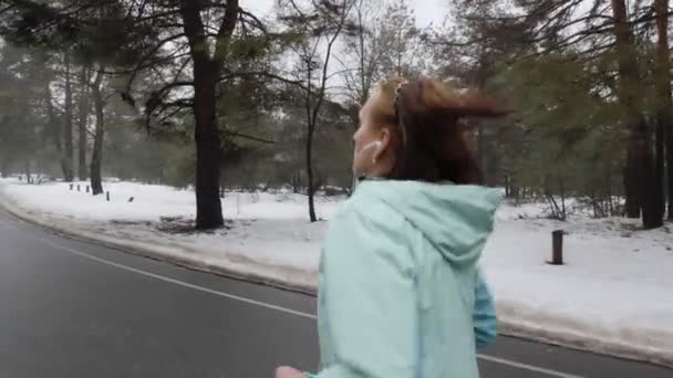 高齢白人女性は、ヘッドフォンで冬に雪の公園で実行しています。バックサイドフォローショット。スローモーション — ストック動画