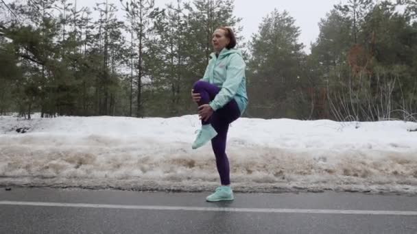 年上の白人女性は雪の冬の公園で実行する前に、ストレッチを温めます。サイドショット. — ストック動画
