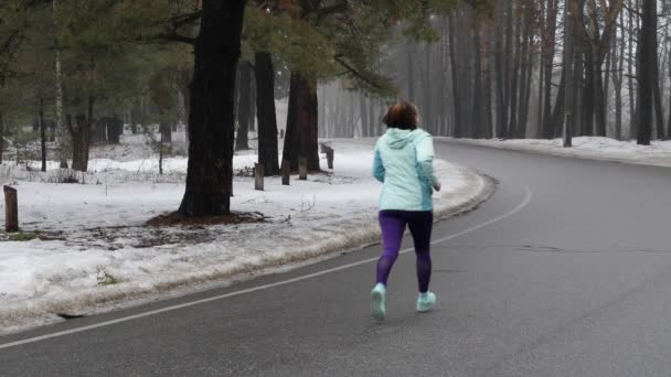Старшая белая женщина бегает в снежном парке зимой в наушниках. Задний статический выстрел. Медленное движение — стоковое видео