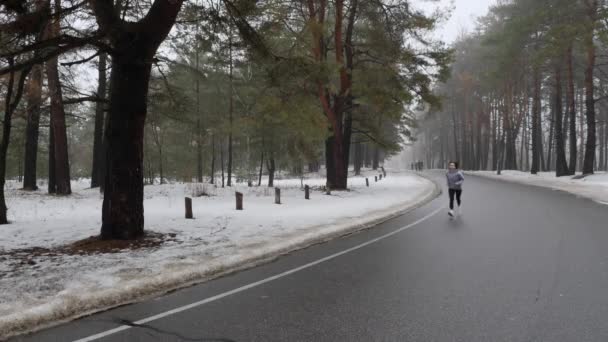 Unga attraktiva kaukasiska flicka som kör i den snöiga parken på vintern med hörlurar. Främre statiska skott. Slow motion — Stockvideo