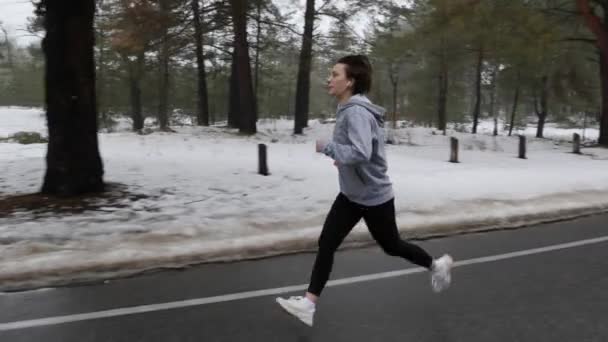 Unga attraktiva kaukasiska flicka som kör i den snöiga parken på vintern med hörlurar. Fram sidan följer skott. Slow Motion (långsam) — Stockvideo