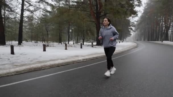 Mladá přitažlivá Kavkazská dívka, která v zimě běží v zasněženém parku se sluchátky. První výstřel. Zpomaleně — Stock video