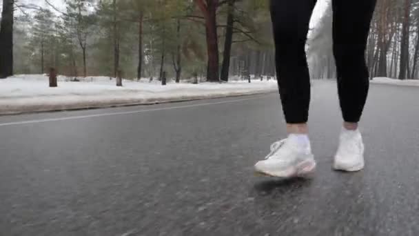 Νεαρό ελκυστικό καυκάσιο κορίτσι τρέχει στο χιονισμένο πάρκο το χειμώνα με ακουστικά. Κοντινό πόδι εμπρός, ακολουθήστε shot. — Αρχείο Βίντεο