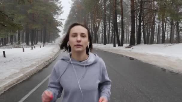 Jonge aantrekkelijke Kaukasische meisje loopt in het besneeuwde Park in de winter met een koptelefoon. Close-up voorkant Volg schot. Slow Motion — Stockvideo