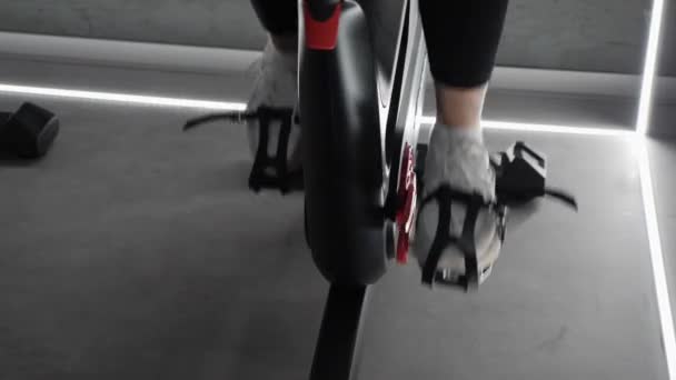 フィットの女の子のクローズアップは、静止バイクでのカーディオサイクリングスピニングワークアウトを行います。スピニング自転車をペダルで閉める — ストック動画