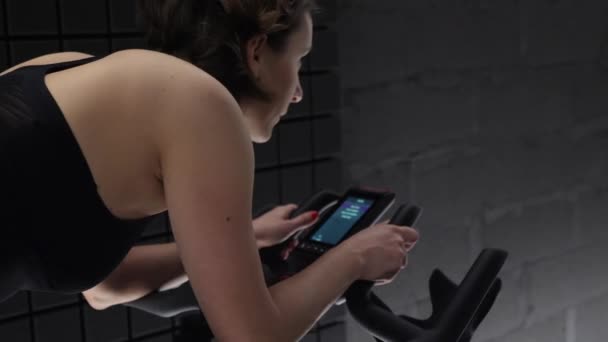 Terug zijaanzicht van mooie aantrekkelijke jonge vrouw genieten van haar workout op hometrainer met computer. Mooi meisje fietsen workout. — Stockvideo