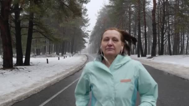 Vieille femme caucasienne âgée courant dans le parc enneigé en hiver avec écouteurs. Fermer avant suivre la prise de vue. Mouvement lent — Video