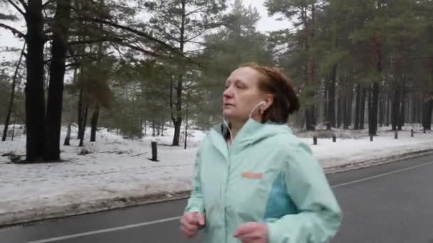 Senior gamla kaukasiska kvinna som kör i den snöiga parken på vintern med hörlurar. Närbild främre följa skott. Slow motion — Stockvideo