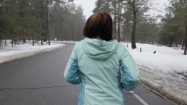 Senior gamla kaukasiska kvinna som kör i den snöiga parken på vintern med hörlurar. Tillbaka följa skott. Slow Motion (långsam) — Stockvideo