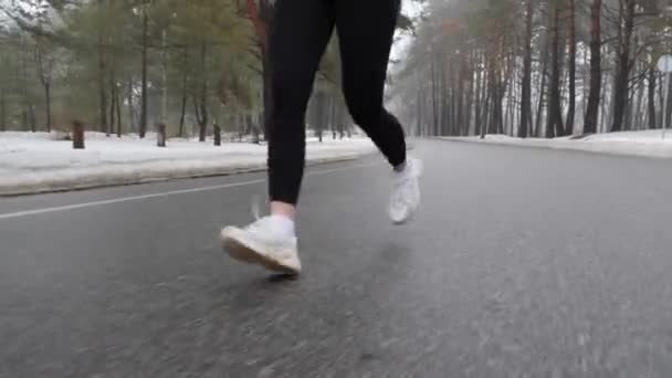 Joven atractiva chica caucásica corriendo en el parque nevado en invierno con auriculares. Primer plano piernas Frente seguir tiro . — Vídeos de Stock