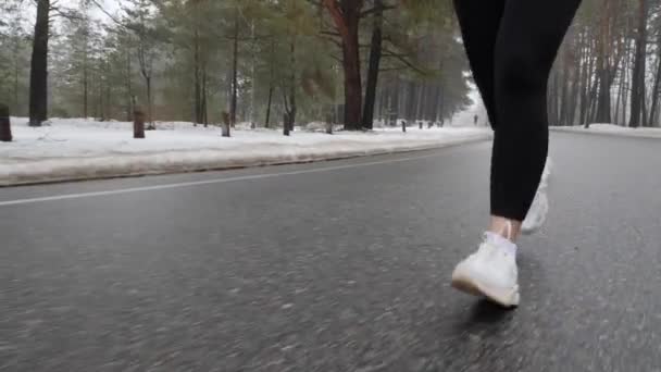 Jovem menina caucasiana atraente correndo no parque nevado no inverno com fones de ouvido. Feche as pernas Frente siga tiro. Movimento lento — Vídeo de Stock
