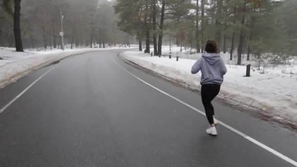 Joven atractiva chica caucásica corriendo en el parque nevado en invierno con auriculares. Volver seguir tiro . — Vídeo de stock