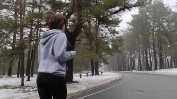 Unga attraktiva kaukasiska flicka som kör i den snöiga parken på vintern med hörlurar. Tillbaka Följ shot. — Stockvideo