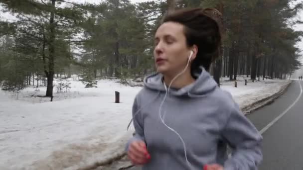 Giovane ragazza caucasica attraente in esecuzione nel parco innevato in inverno con le cuffie. Avvicinati al front follow shot. Rallentatore — Video Stock