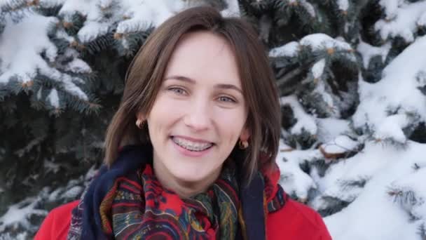 Красивая молодая кавказка, завернутая в тёплое красное пальто с шарфом, улыбающаяся в камеру с зубными брекетами — стоковое видео
