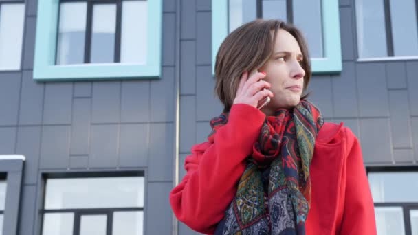 Mooie jonge zakelijke vrouw praten via de telefoon tegen de complexe van de moderne kantoorgebouw in rode jas en sjaal — Stockvideo