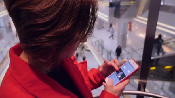 Joven atractiva dama de negocios caucásica en abrigo rojo usando su teléfono en un ascensor — Vídeos de Stock