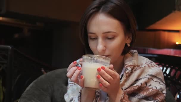 年轻而有魅力的女人正在餐馆里喝咖啡茶。她的肩膀上覆盖着一条温暖的针织围巾 — 图库视频影像