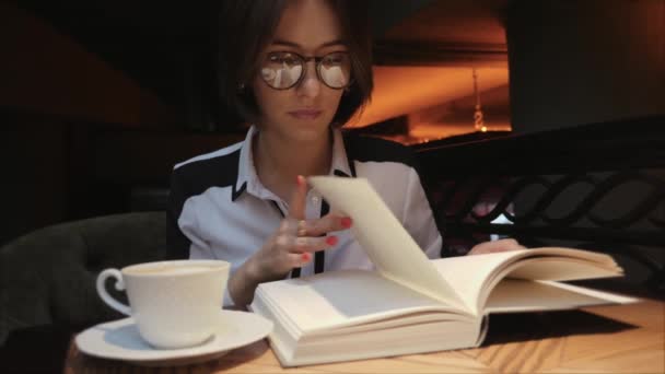 Νεαρή γυναίκα που ανοίγει και διαβάζει ένα βιβλίο πίνοντας καφέ σε φιλόξενο καφέ — Αρχείο Βίντεο