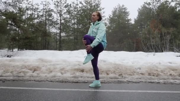 La anciana caucásica se calienta estirándose antes de correr en el nevado parque de invierno. Tiro lateral . — Vídeo de stock