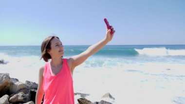 Kayalık plajda ayakta iken telefon selfie fotoğrafları yapıyor ile genç güzel beyaz kız. Dalgalar kayaların üzerine dalıyor. Yavaş hareket