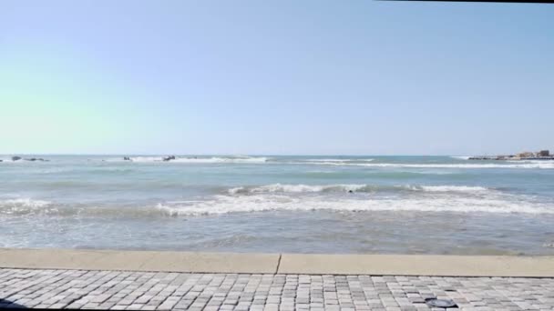 Молодая красивая кавказская девушка с телефоном делает селфи фотографии, стоя на скалистом пляже. Волны брызгают по скалам — стоковое видео