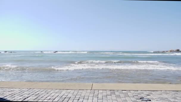Молодая красивая кавказская девушка с телефоном делает селфи фотографии, стоя на скалистом пляже. Волны брызгают по скалам — стоковое видео