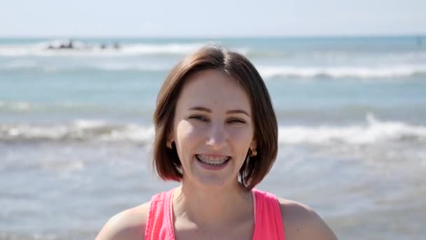 Joven chica en forma hermosa sonriendo con frenillos en la playa con el océano ondulado — Vídeos de Stock