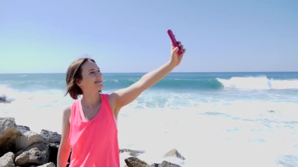 年轻美丽的高加索女孩与电话做自拍照片, 而站在岩石海滩上。海浪在岩石上飞溅 — 图库视频影像
