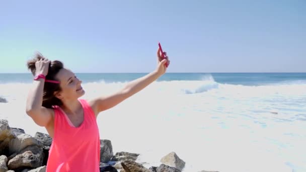 Joven hermosa chica caucásica con teléfono haciendo fotos selfie mientras está de pie en la playa rocosa tocando su cabello y sonriendo — Vídeos de Stock