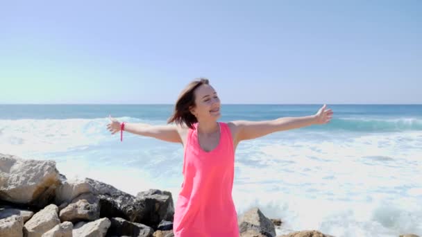 Jeune fille heureuse souriante écartant les bras tout en se tenant sur la plage rocheuse. Des vagues frappent des rochers. Des éclaboussures. Mouvement lent — Video