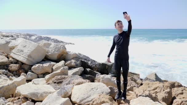 Giovane uomo felice sport sottile in piedi sulla spiaggia rocciosa e prendere selfie mentre l'acqua schizza contro le scogliere. Rallentatore — Video Stock