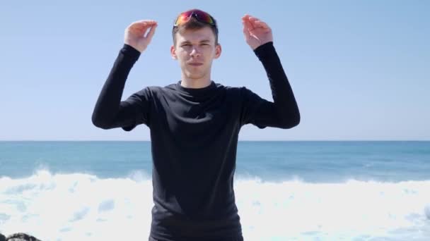 Młody pewny siebie sportowcy stawia na swoje okulary stojące na plaży z falami za. Słoneczny dzień — Wideo stockowe