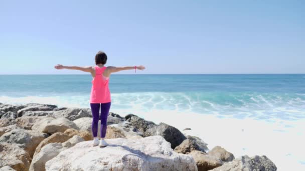 Young caucasian brunette girl standing on a rocky beach with arms open wide while strong waves hitting the cliffs and water splashing. Sunny windy day. Slow Motion — Stock Video