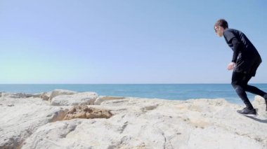 Plajda kayalar üzerinde çalışan siyah genç sportif uygun beyaz adam. Dalgalar güneşli bir rüzgarlı gün üzerinde sıçrama.