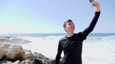 Siyah genç sportif uygun beyaz adam selfie 'ler alarak ve dalgalar vurmak ve su sıçraması ile kayalık plajda ayakta gülümseyen. Yavaş hareket
