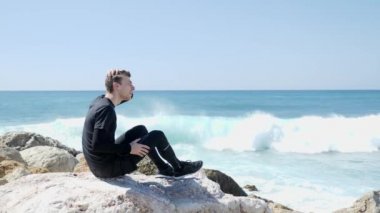 Genç beyaz adam saçlarında parmaklarıyla kayalar üzerinde oturuyor. Kayalık plaja su sıçraması ile çarpan güçlü okyanus dalgaları. Güneşli bir gün. Yavaş hareket