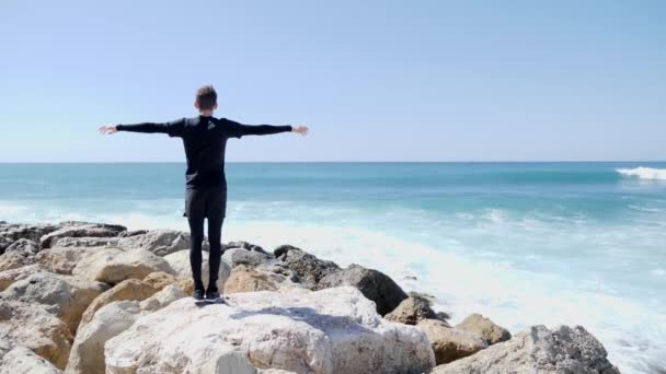 岩のビーチに立っている若い白人男性は、広い波が崖と水しぶきに当たる間、腕を広げています。サニー風の日. — ストック動画