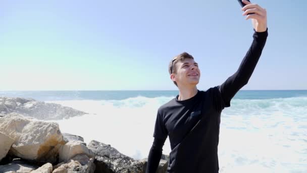 Jeune homme caucasien en forme sportive en noir prendre selfies et sourire tout en se tenant sur la plage rocheuse avec des vagues frappant et éclaboussures d'eau. Mouvement lent — Video