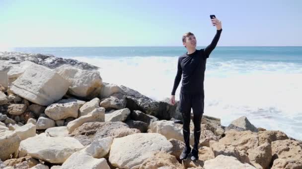 Jeune homme heureux sportif mince debout sur la plage rocheuse prendre selfies et montrant pouces vers le haut tandis que l'eau éclaboussure à nouveau les falaises — Video