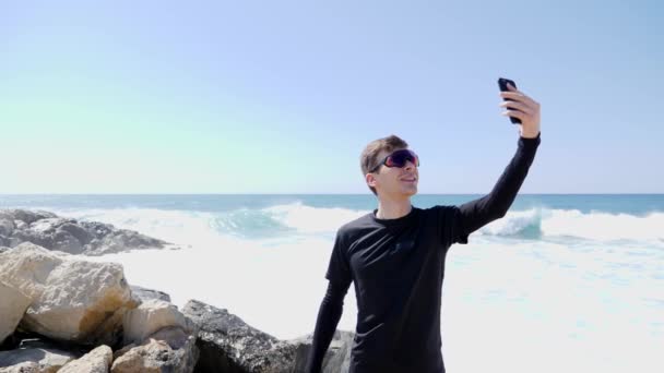 Молодой спортивный белый мужчина в черных очках в солнечных очках держит телефон и делает селфи, стоя на скалистом пляже с ударами волн и брызгами воды. Slow Motion — стоковое видео